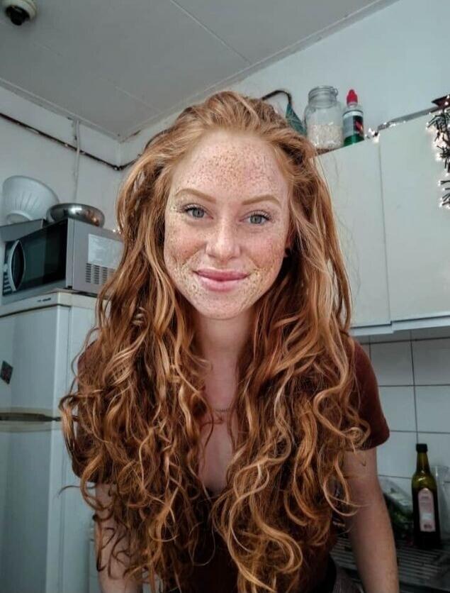 A smiling woman with long curly red hair and freckles in a kitchen.