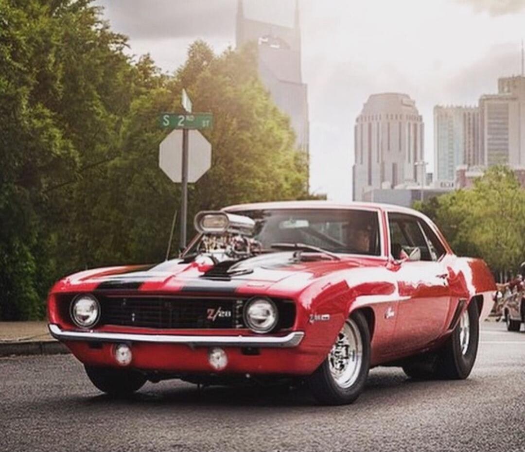 Red vintage muscle car on a city street with a skyline in the background. Street sign reads S 20th.