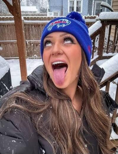 A young woman wearing a blue Buffalo Bills beanie with a pom-pom, outdoors in the snow, looking up with her mouth open and tongue sticking out for a playful selfie. She is on a wooden deck with snow-covered railings and surroundings.