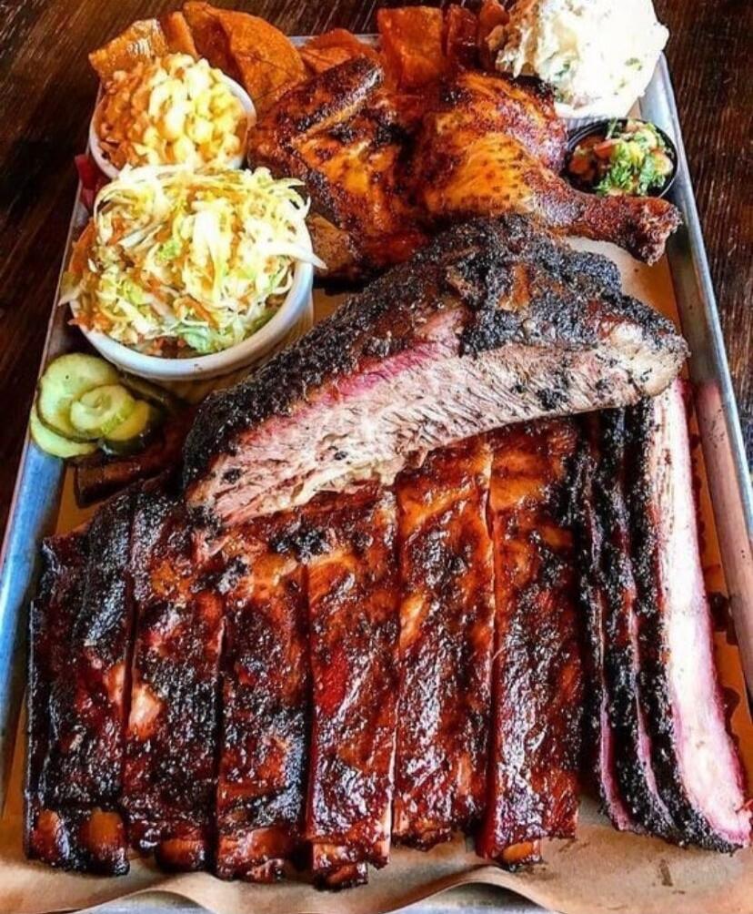 A large platter of barbecue meat, including smoked ribs, sliced brisket, and a roasted half chicken, served with various side dishes such as coleslaw, macaroni salad, and sliced pickles on a metal tray.