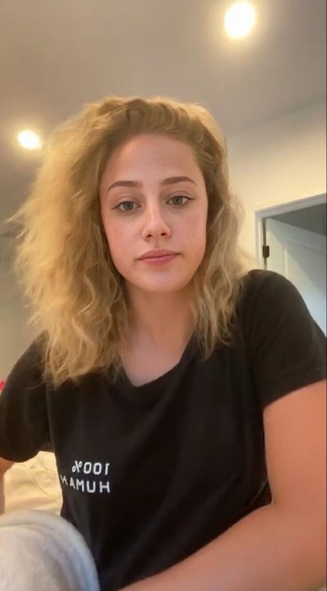 A young woman with blonde, wavy hair, wearing a black t-shirt with the mirrored text '100% HUMAN'. She is looking directly at the camera.