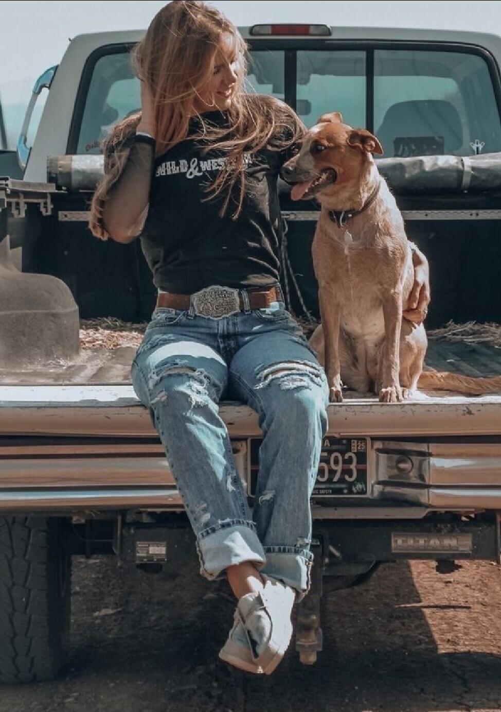 A young woman sits on the tailgate of a pickup truck with a tan dog beside her. She wears a black cropped t-shirt, ripped blue jeans, white sneakers, and a large belt buckle. The dog sits close, looking up at her as she touches her hair.