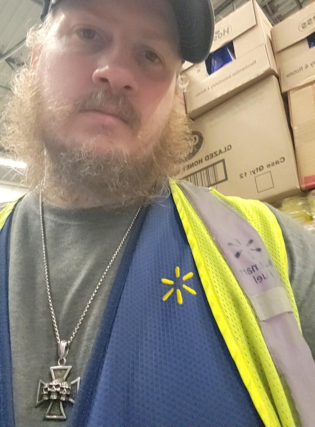 A man with a beard and a cap, wearing a grey t-shirt, a blue Walmart vest with a yellow spark logo, and a yellow high-visibility vest. He has a necklace with a skull-themed cross pendant. In the background, there are stacks of cardboard boxes, some with text like 'CLOSED HOME', 'HO', 'CASE OF 12', '225'.