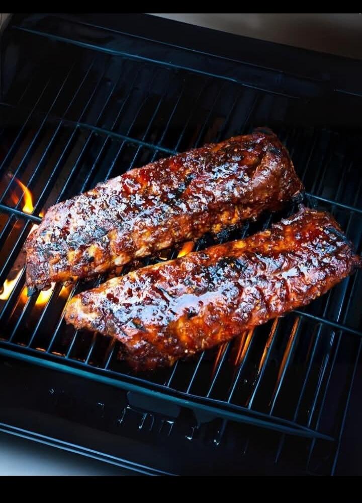 Two racks of ribs are grilling on a barbecue, coated in a shiny, dark glaze. Flames are visible beneath the grill grates, suggesting high heat and a smoky flavor.