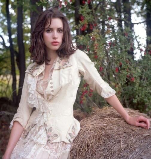 A young woman in a cream vintage lace dress stands leaning on a hay bale in a garden setting.