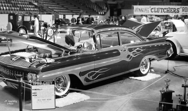 A black and white photograph shows a customized 1959 Chevrolet car on display at an indoor car show. The car features a distinctive flame paint job on its sides. The hood is open, revealing the engine. A sign in front of the car reads '1959 CHEV' and 'WILD PAINT WORK'. In the background, a banner with 'WINNIPEG CLUTCHERS 