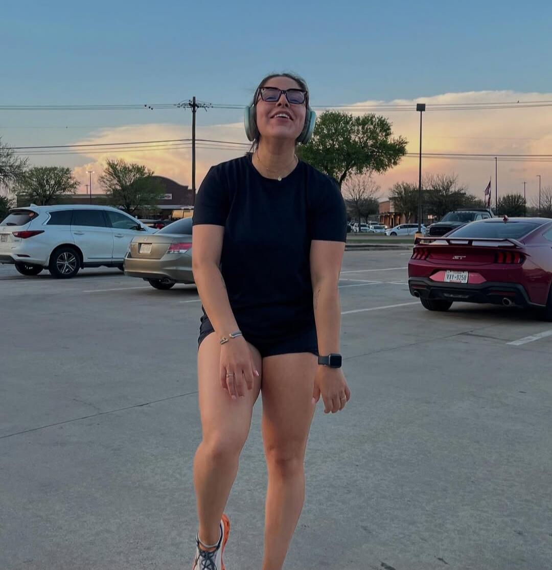 A woman in a black t-shirt and black shorts, wearing glasses and headphones around her neck, smiles happily in a parking lot at sunset. Several cars are visible in the background.