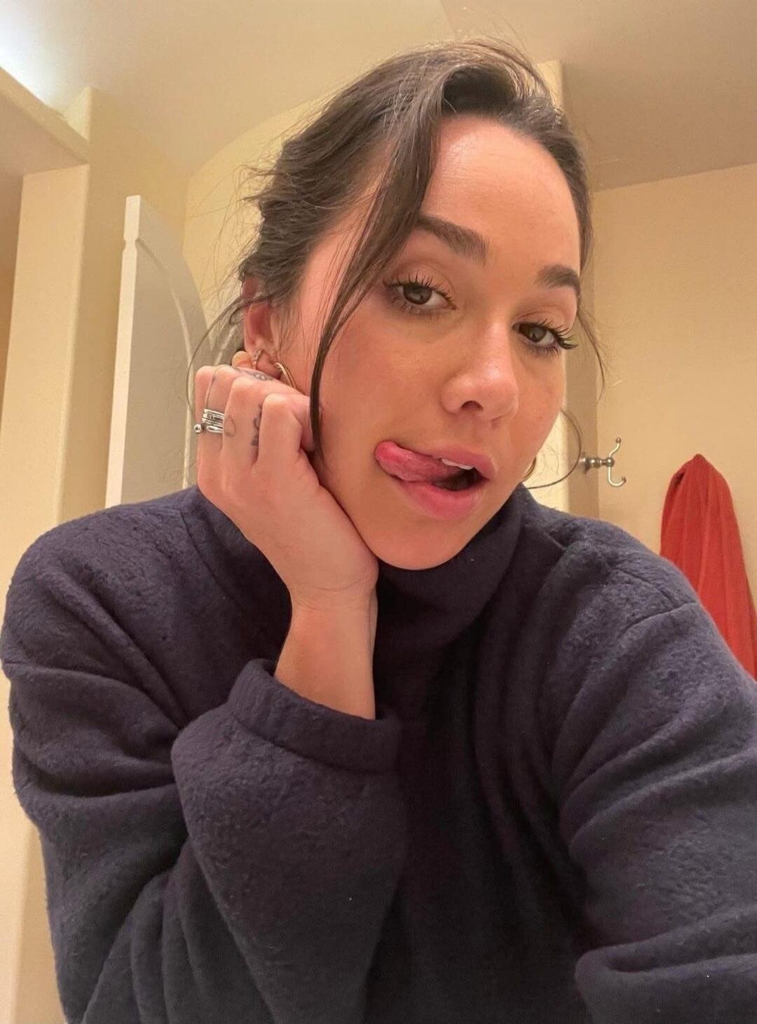 A woman in a dark turtleneck sweater takes a selfie, playfully sticking her tongue out. Her hand is resting on her chin, showing rings on her fingers. The background is a light-colored room, possibly a bathroom, with a red towel visible.