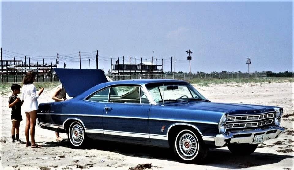 A blue vintage car parked on a sandy beach with its hood open. A group of people (two women and two children) are standing near the car.