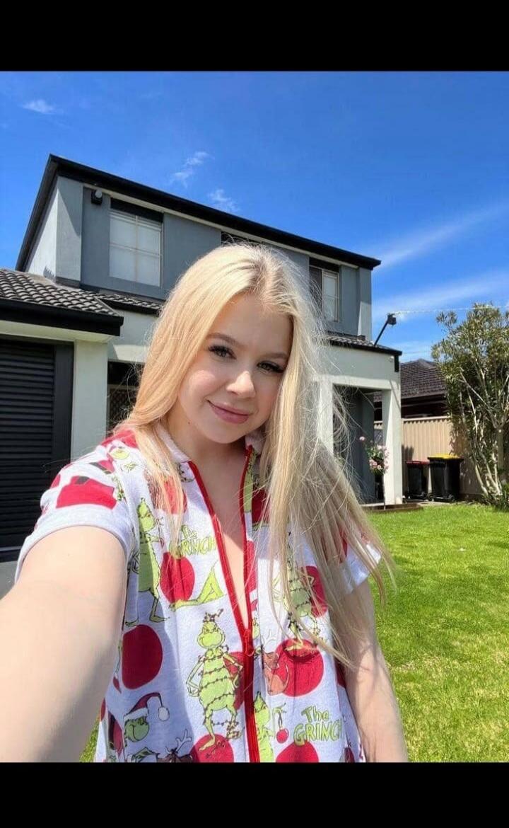 A young blonde woman takes a selfie outdoors in front of a house. She is wearing a white top with 'The Grinch' and Christmas ornament patterns. The sky is blue and clear.