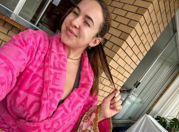 A woman in a pink bathrobe is taking a selfie, winking and smiling at the camera. She is holding a strand of her hair. A dark top is visible under the bathrobe.