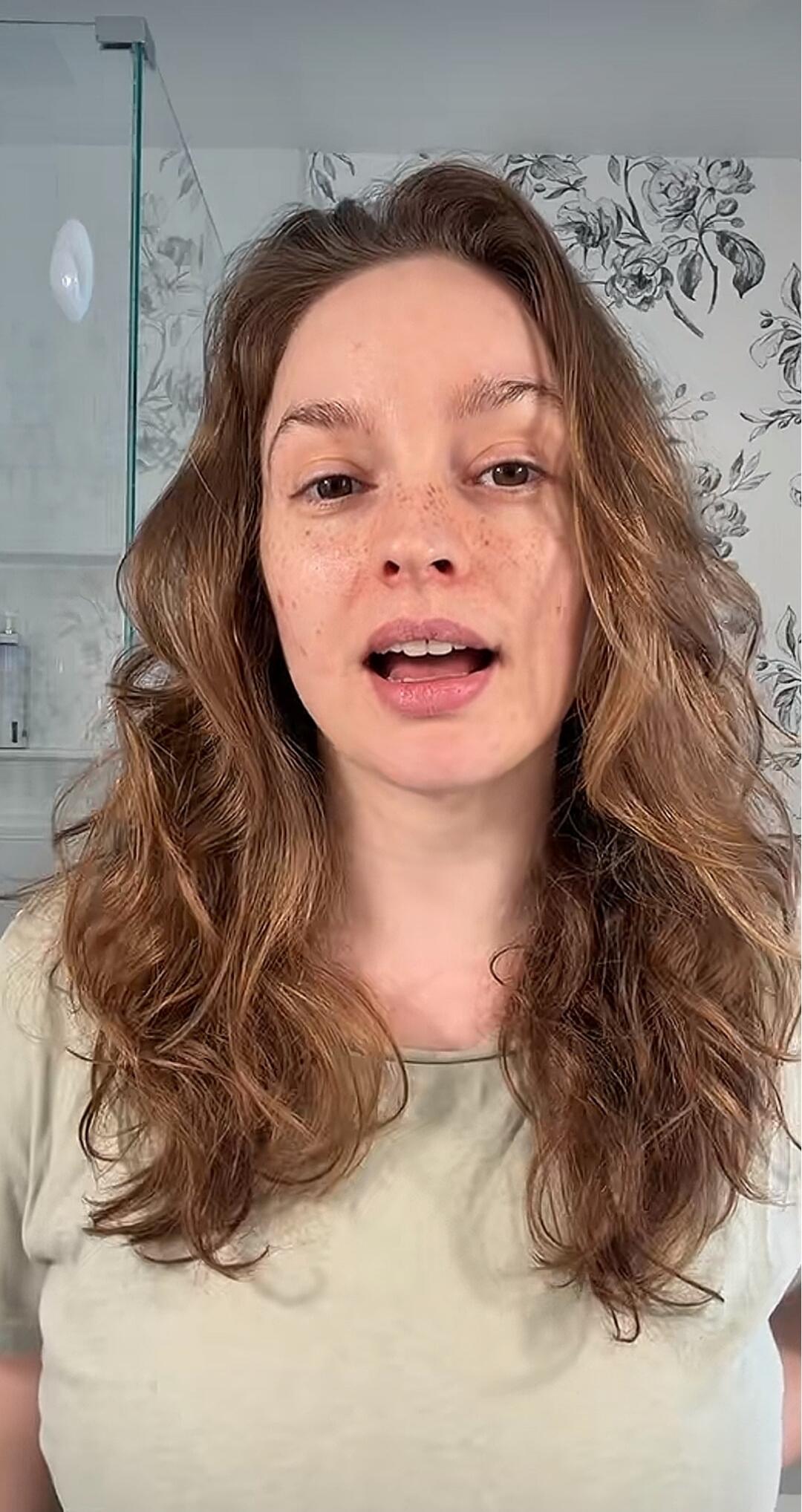 A woman with long wavy brown hair and freckles is speaking directly to the camera. She is wearing a light green t-shirt. Her mouth is open, suggesting she is mid-sentence or explaining something. The background appears to be a bathroom with a glass shower partition and floral wallpaper.