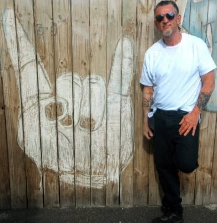A man in sunglasses and a white t-shirt stands next to a wooden fence with a large drawing of a hand making the 'rock on' gesture.