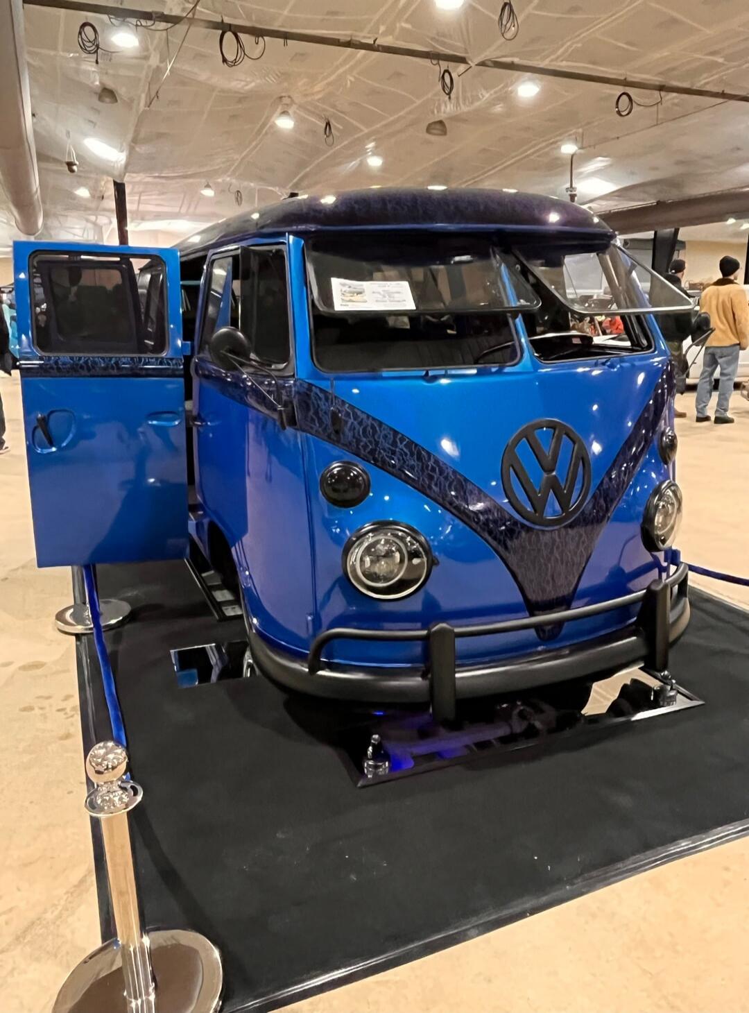 Blue vintage Volkswagen van on display at an indoor exhibit. The classic split-window VW bus is painted bright blue with a black diagonal stripe and is surrounded by a small barrier setup.