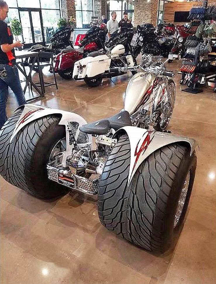 Session ID: 1064876. A white/silver three-wheeled motorcycle with oversized tires on display inside a showroom, surrounded by other motorcycles.