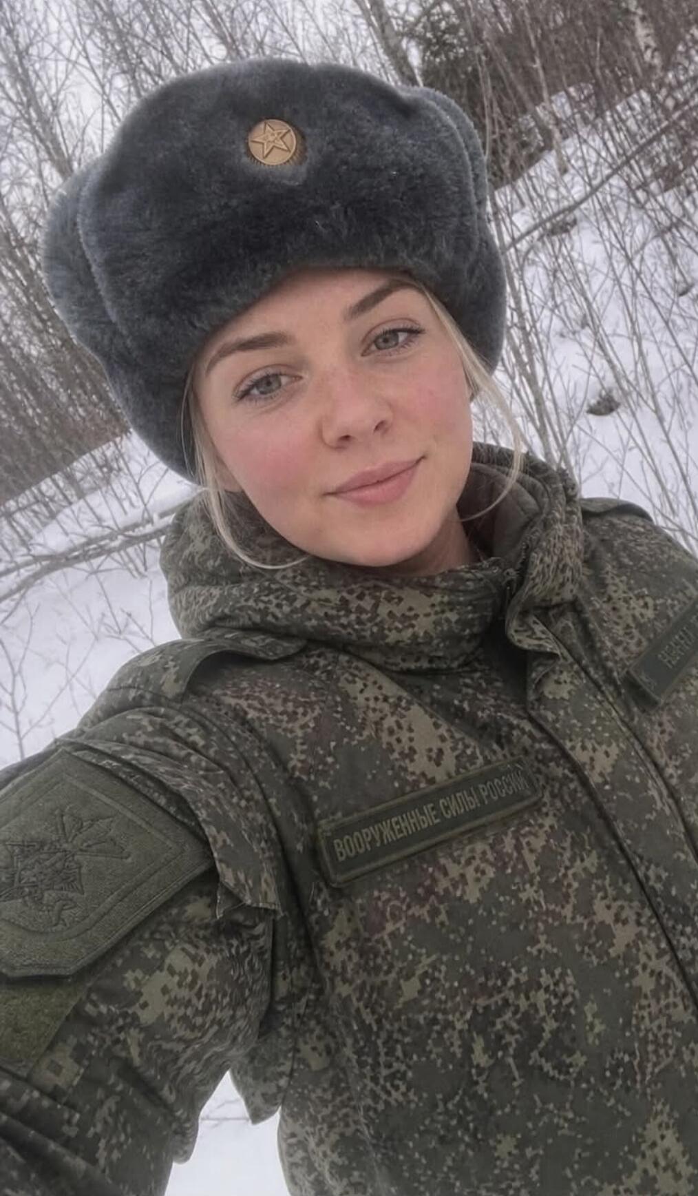A young woman in a Russian military uniform and an ushanka hat, smiling. The uniform has a patch that reads 'ВООРУЖЕННЫЕ СИЛЫ РОССИИ'.
