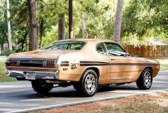 A vintage gold or bronze colored car, possibly a Dodge Demon or Dart, is parked on the side of a road. The car has black racing stripes on its sides. The license plate reads 'TEXAS 9DY3723'. Trees and foliage are visible in the background. The tires have 'BFGoodrich' written on them.