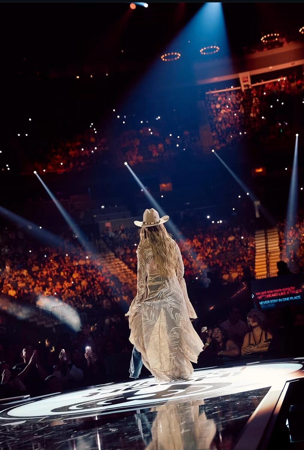 we got one more song for WITH HEART LIKE A TRUCK NEVER STAY IN ONE PLACE SO LONG. A performer on stage, wearing a cowboy hat and a flowing sheer top, faces a large audience in a concert venue. Bright spotlights illuminate the stage, and the crowd holds up their phones.
