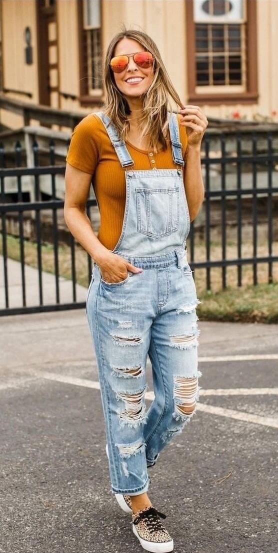 A woman wearing light-wash ripped denim overalls and an orange-brown short-sleeve top, orange-tinted sunglasses, standing outdoors with a smile.