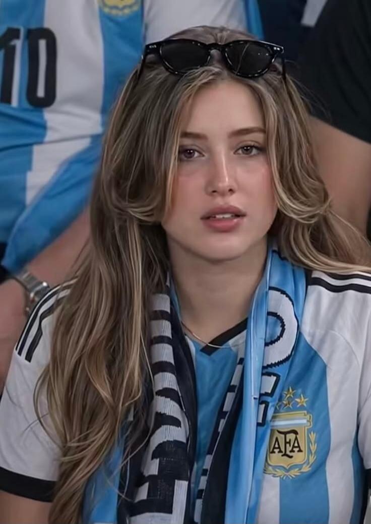 A young woman wearing an Argentina national football team jersey with the 'AFA' logo and a scarf, with the number '10' visible on a jersey in the background. She has sunglasses on her head.