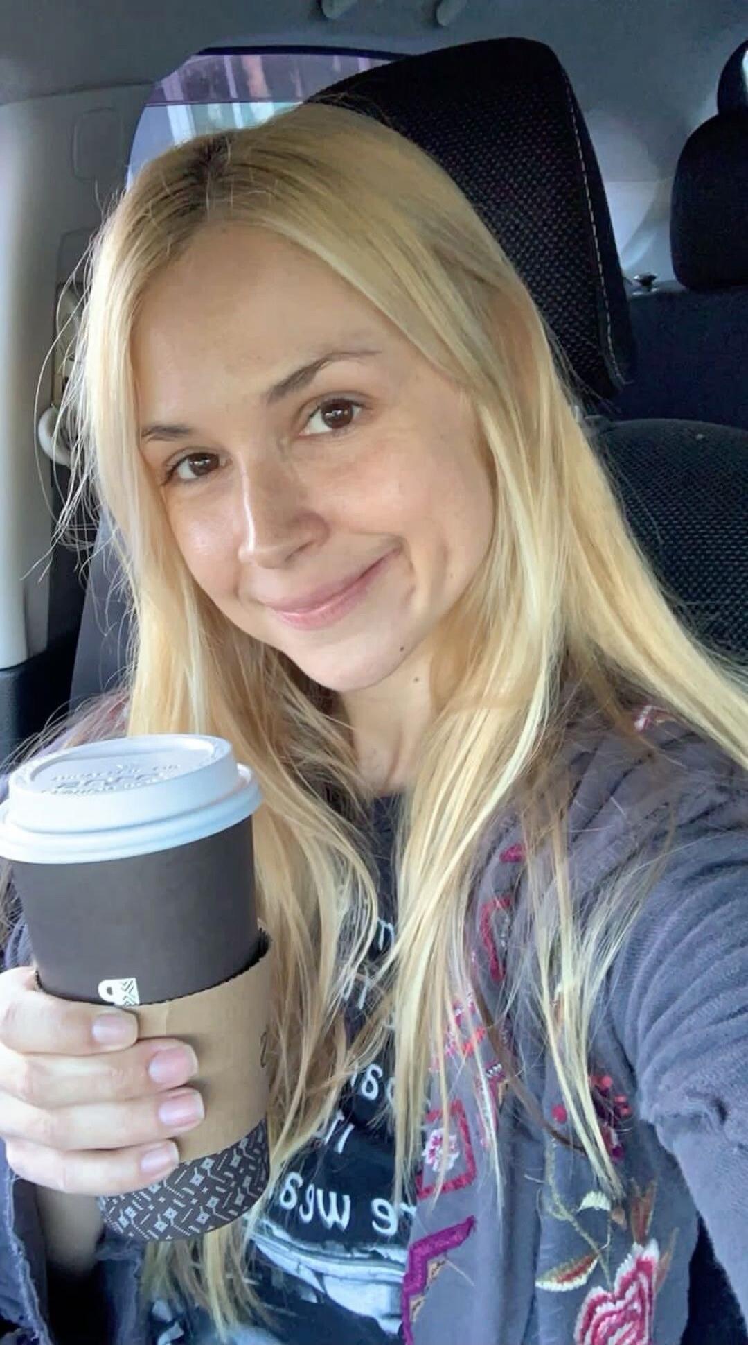 A blonde woman in a car takes a selfie, holding a coffee cup. She is smiling. Text on her shirt appears to be inverted, possibly '69w 9'. A small coffee cup icon is visible on the cup sleeve.
