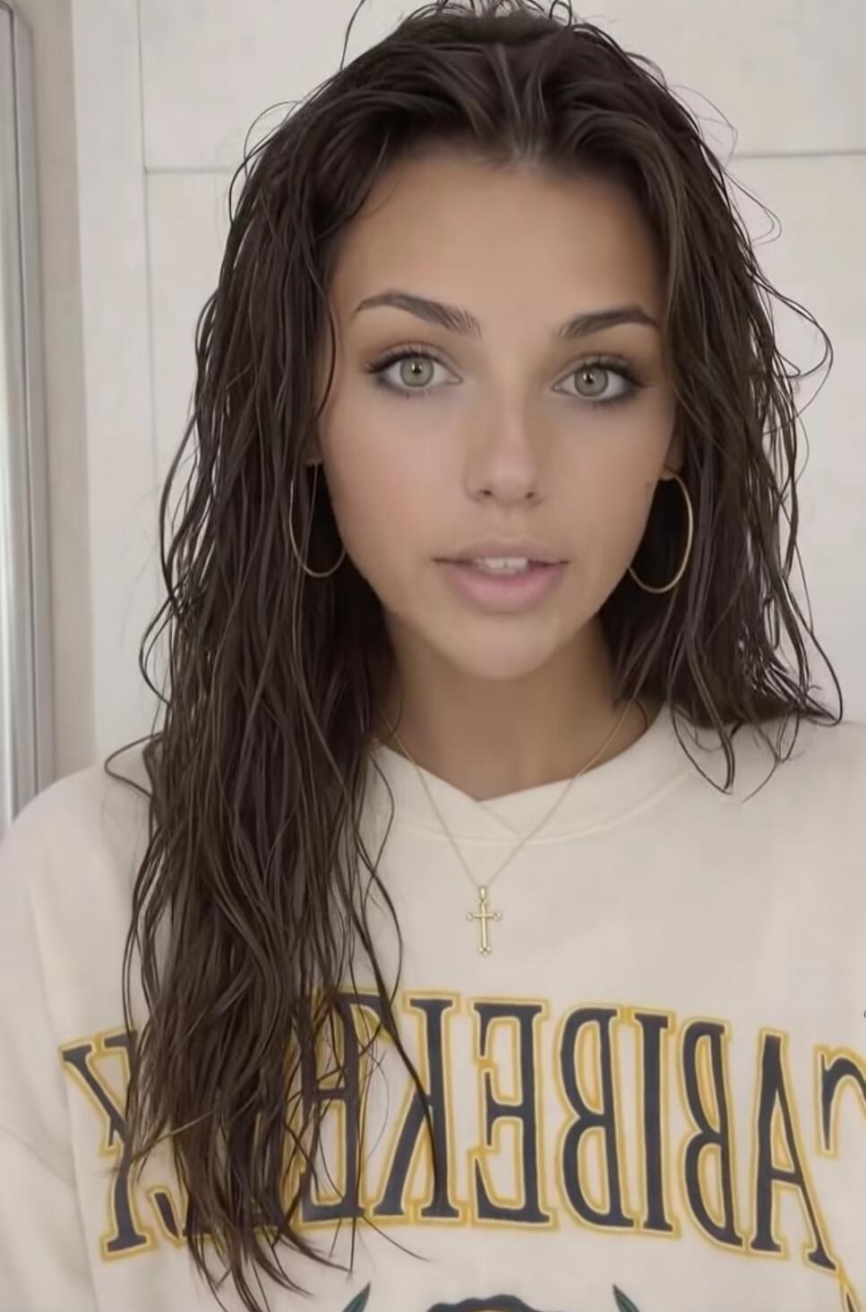 A young woman with wet, dark hair and striking green eyes, wearing a light-colored sweatshirt with the word 'BERKELEY' printed on it (reversed) and a cross necklace. She is also wearing hoop earrings and looking directly at the camera.