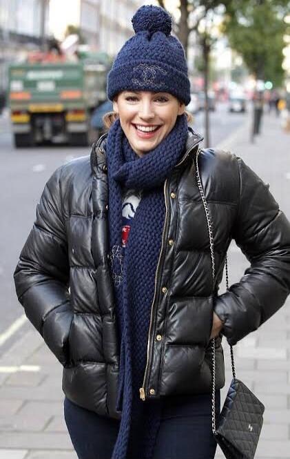 A smiling woman on a city street wearing a black puffer jacket, blue scarf and beanie.
