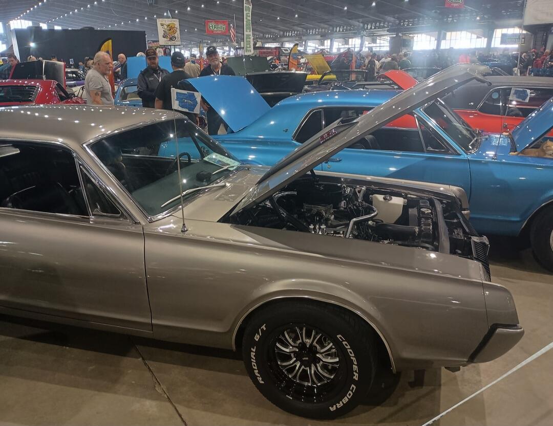 An indoor car show featuring classic and custom automobiles. The foreground shows a silver/grey muscle car with its hood open, revealing the engine. In the background, other vintage cars and people are visible, walking around and admiring the vehicles.