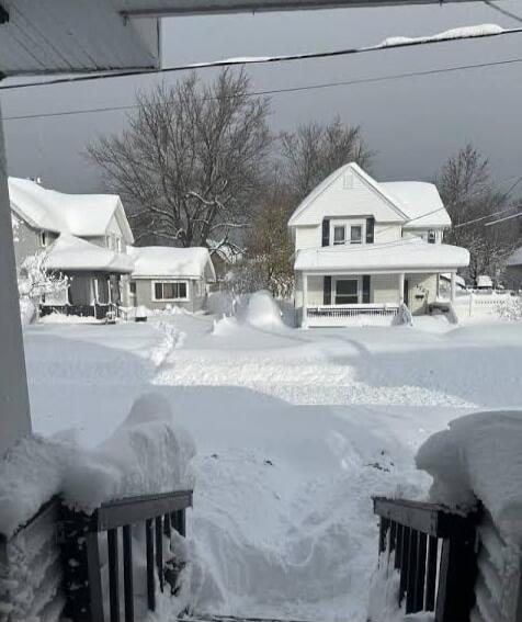 A snowy suburban street with heavy snow covering houses, trees, and fences. Large snowdrifts on sidewalks and driveways, snow piled high around yards.