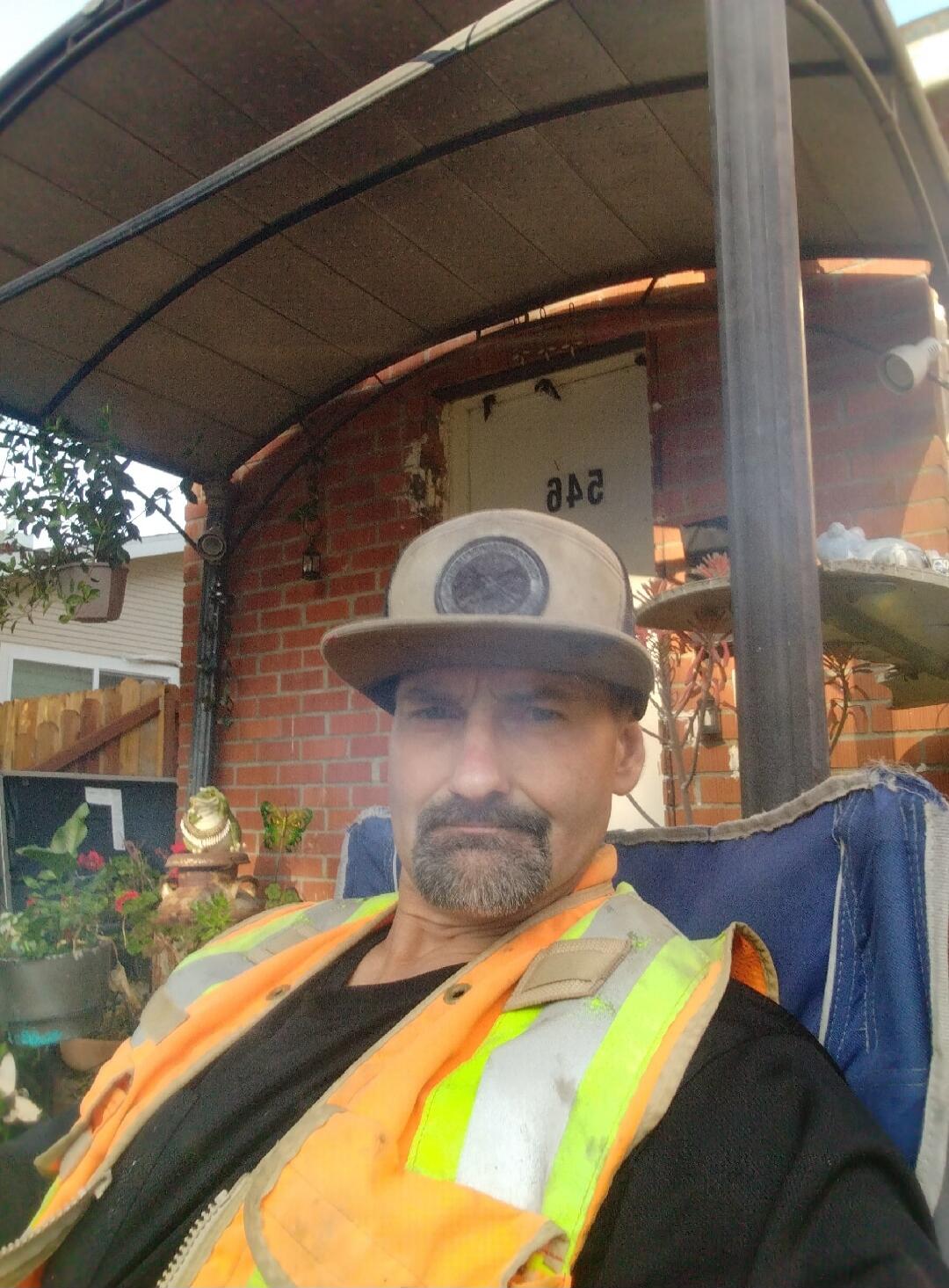 A man wearing a beige cap and an orange high-visibility vest is sitting in a chair. In the background, there's a brick wall, a white door with the number '242' (reversed), and potted plants.