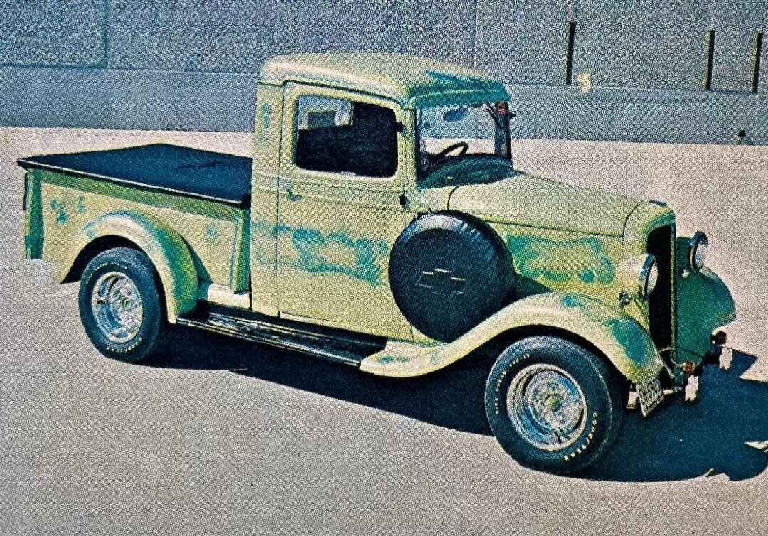 This is an image of a vintage light green pickup truck with custom paintwork. The truck has a spare tire mounted on the front fender and a black tonneau cover over the bed. The paint has a faded, airbrushed design that appears to be flames or clouds. The truck is parked on an asphalt surface with a textured wall in the background.