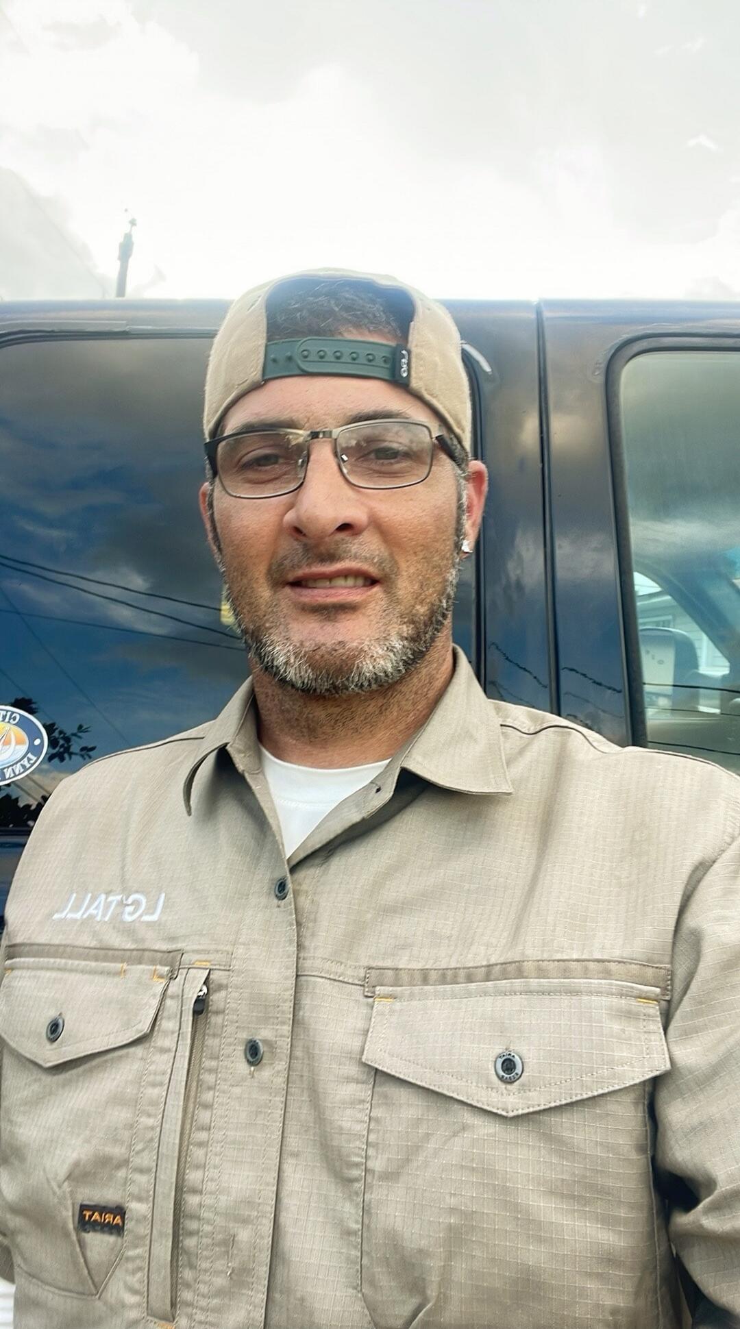 A man in a cap, glasses, and a work shirt with a beard is taking a selfie. Text visible on the shirt includes 'LATTAL' (reversed) and 'TAIRA'. A sticker on the vehicle behind him shows 'CITY OF' (reversed).
