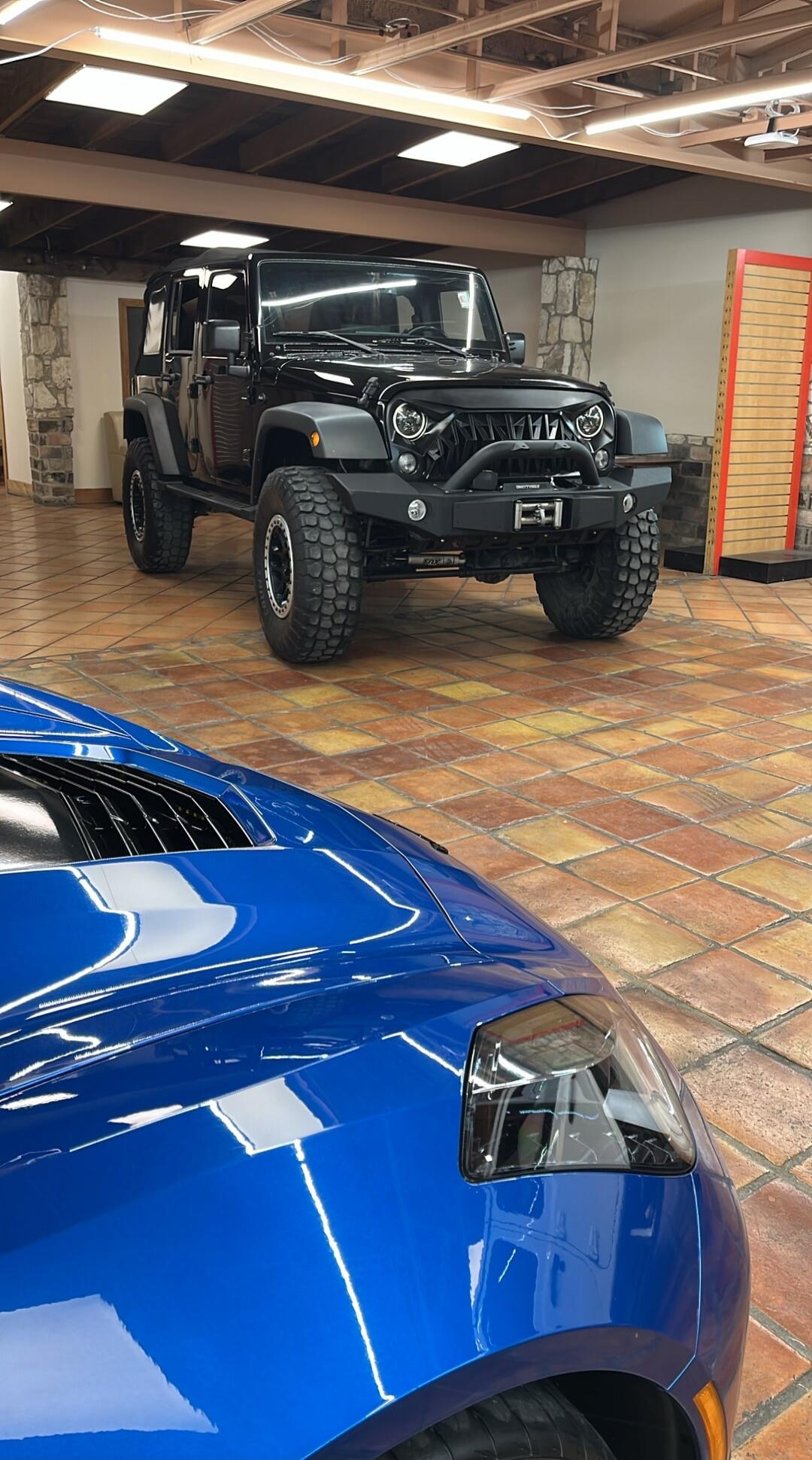 Black modified Jeep Wrangler with large off-road tires in a showroom, with a blue sports car in the foreground.