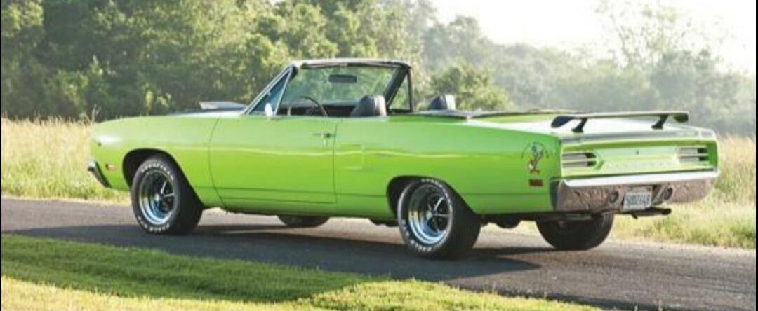 A bright green classic convertible car is parked on an asphalt road next to a grassy field with trees in the background.