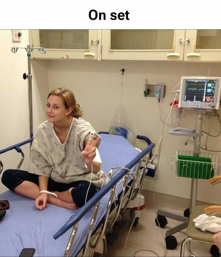 On set. A young woman in a hospital gown is sitting on a hospital bed, making a peace sign with her hand. She has a pulse oximeter on her finger and an ID band on her wrist. Hospital equipment is visible in the background.