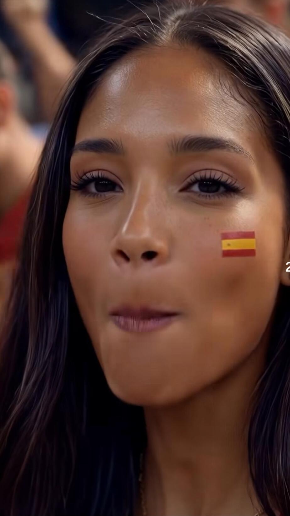 A close-up of a young woman with long dark hair, looking at the camera. She has a Spanish flag painted on her cheek and her lips are slightly pursed. She appears to be a fan at a sporting event.