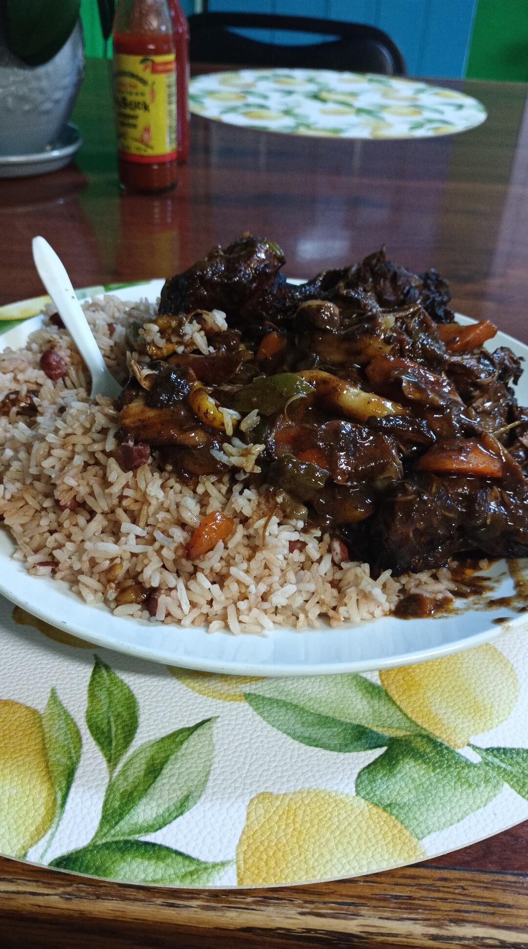 Plate of rice with braised beef and vegetables, served with a dark sauce. A bottle of hot sauce sits on the table in the background.