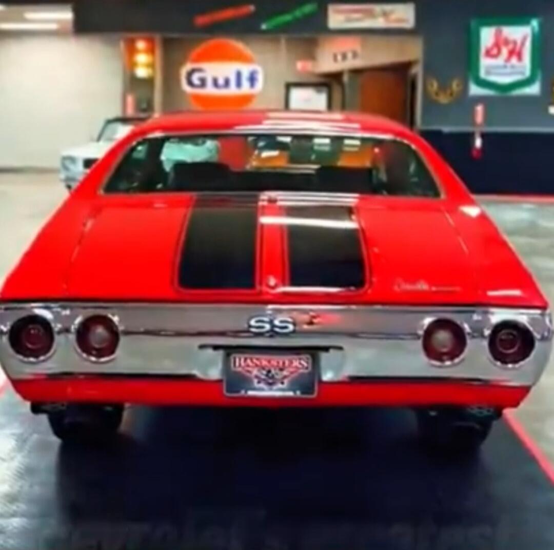 A red classic muscle car, a Chevrolet Chevelle SS, with black racing stripes on the trunk. The car is parked indoors, possibly in a showroom or garage, with a 'Gulf' sign visible in the background. The license plate reads 'HANKSTERS'.