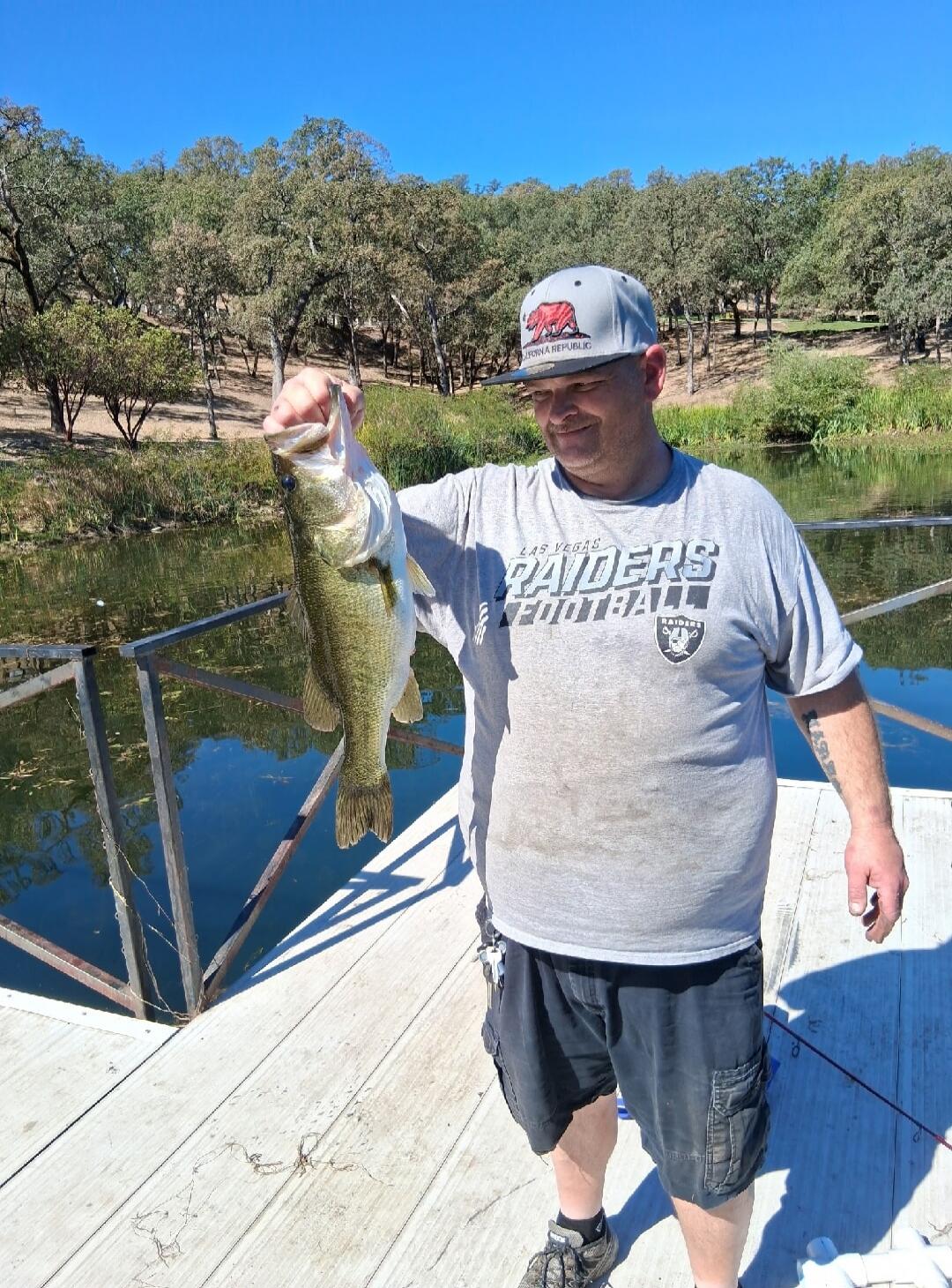 A man holding a large bass fish on a wooden dock. He is wearing a grey t-shirt with 'LAS VEGAS RAIDERS FOOTBALL' printed on it and a grey cap with a bear logo and 'CALIFORNIA REPUBLIC'. The background shows a calm body of water surrounded by trees under a clear blue sky.