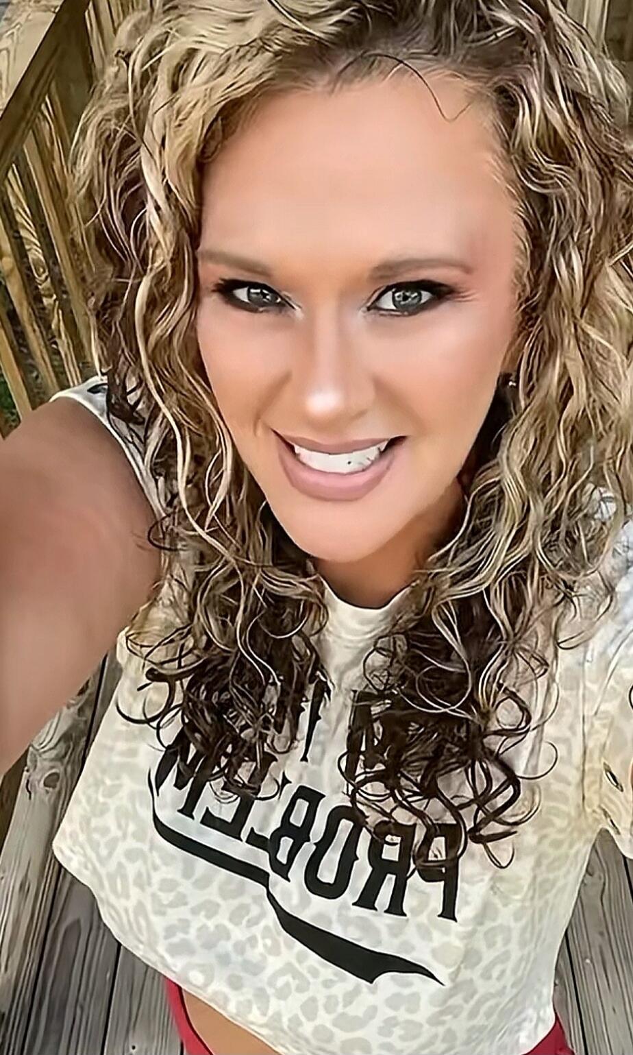 A woman with curly blonde hair smiles at the camera, wearing a leopard print crop top with the word 'PROBLEM' printed on it (reversed).