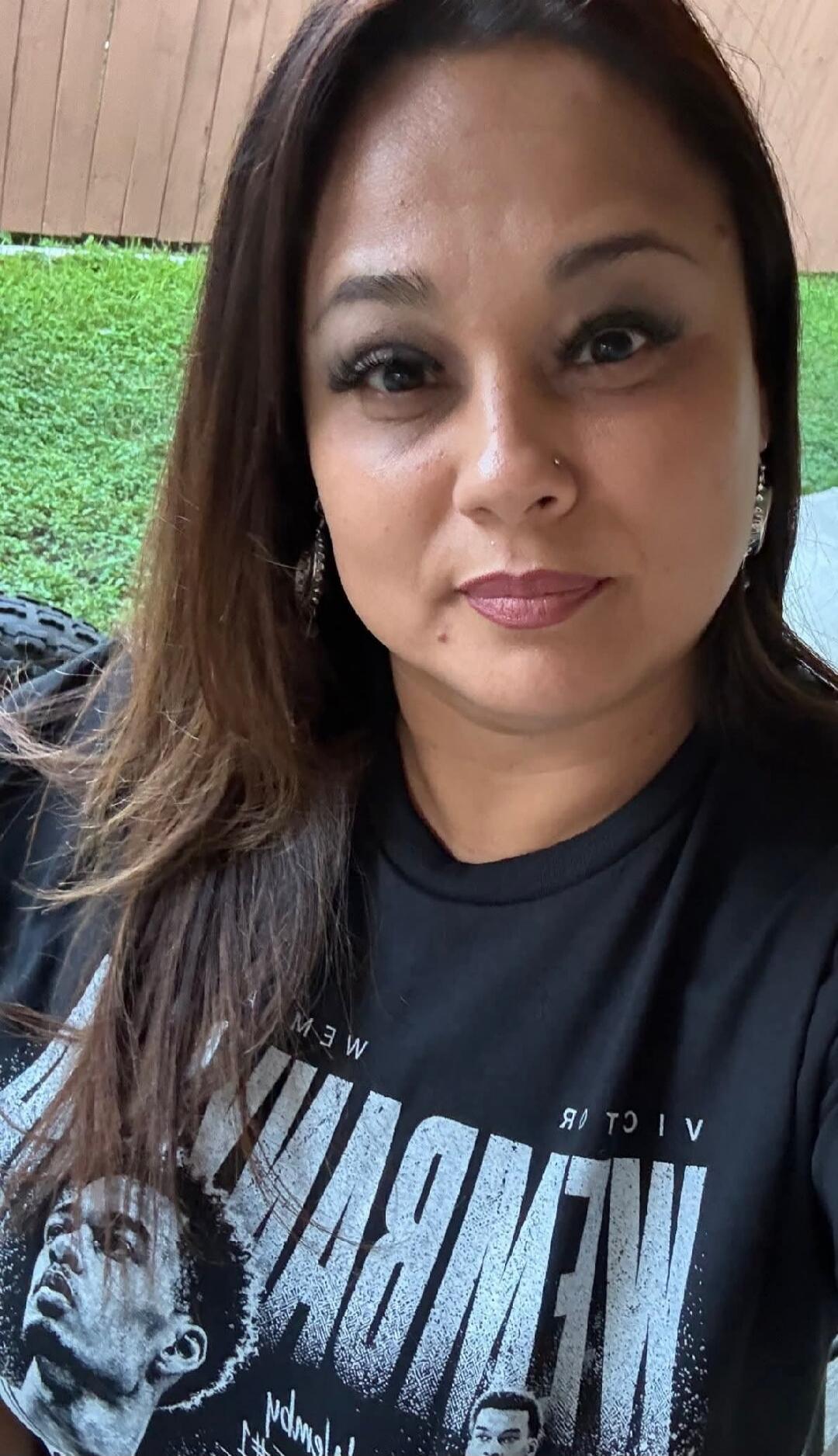 A woman takes a selfie, looking directly at the camera. She has long dark hair and is wearing a black t-shirt with some mirrored white text and images.