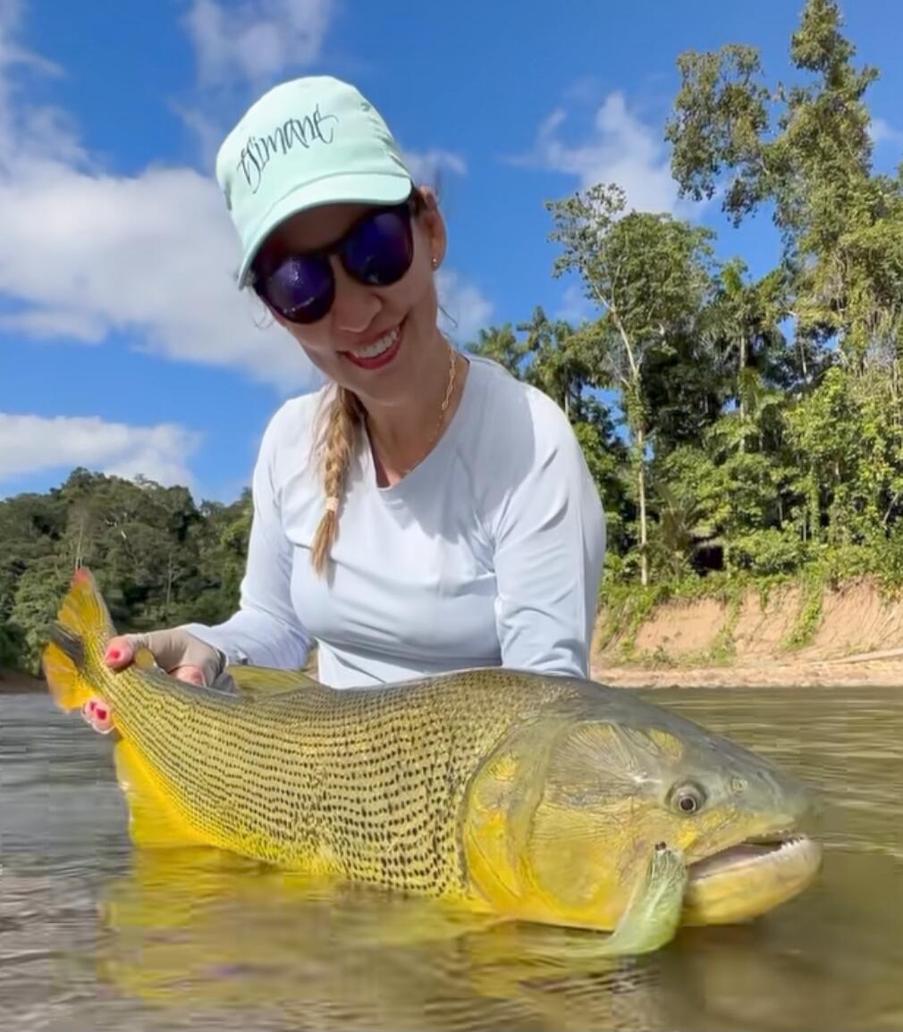 A woman in sunglasses and a cap holds a large yellow-green fish in shallow water.