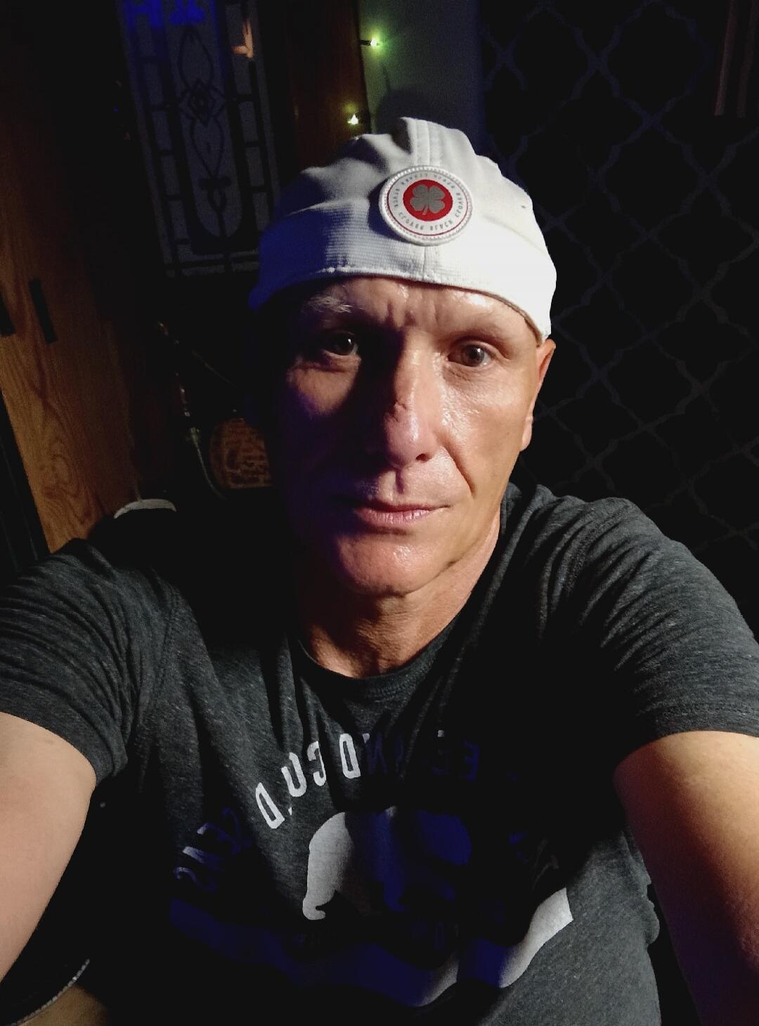 A man wearing a white cap with a red clover logo and a dark grey t-shirt. The text 'CLOVER' is visible on the cap. The text on the t-shirt is inverted and difficult to read.