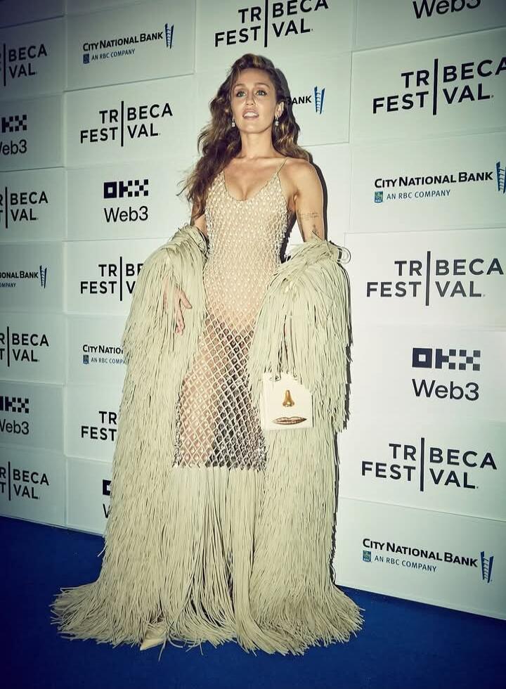 TRIBECA FESTIVAL (logos visible: City National Bank, Web3, NBC) on backdrop. Woman on blue carpet wearing a long fringed cream/beige dress with a sheer inner layer and large fringe coat.