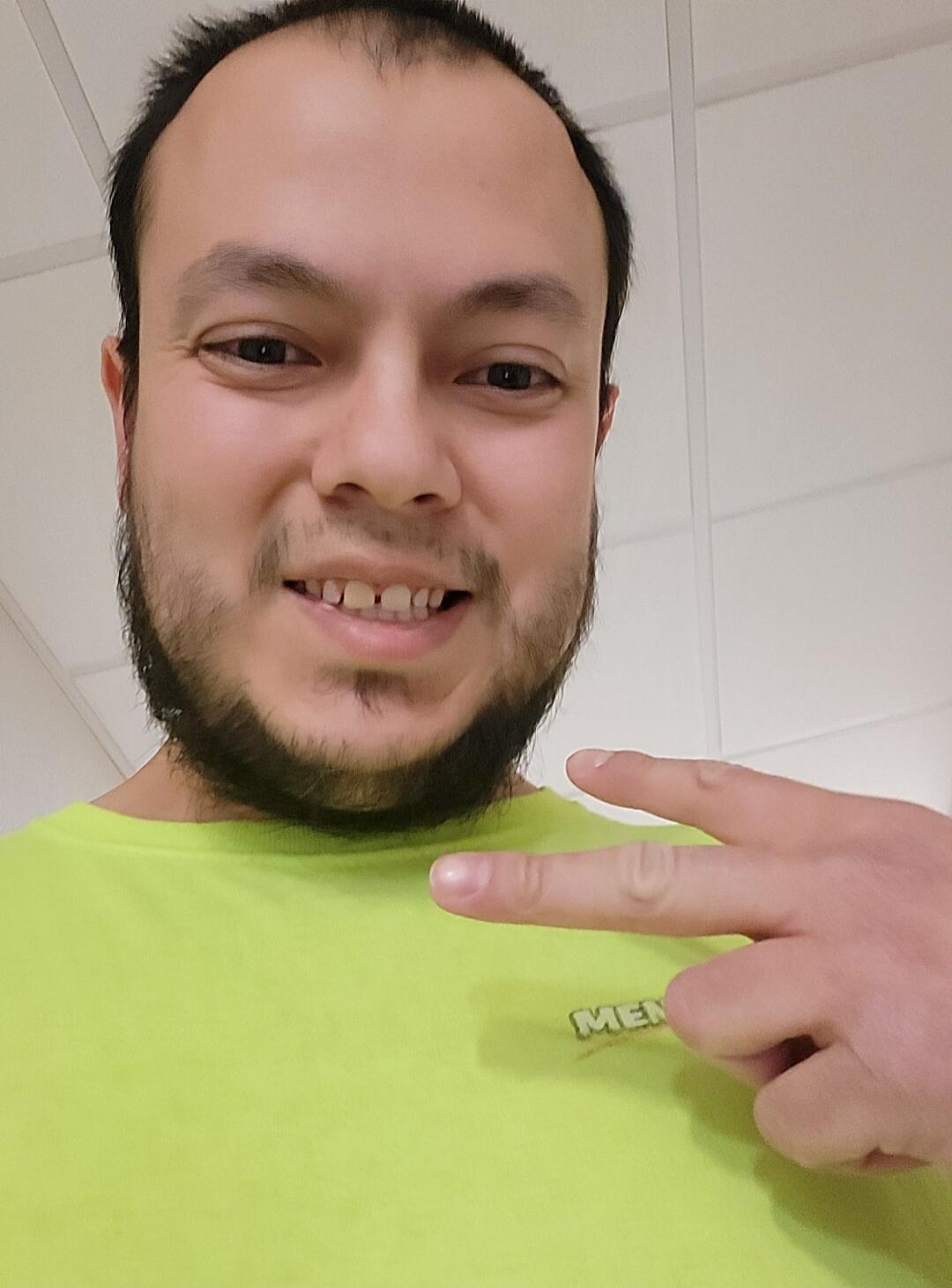 A man with a beard and a bright green t-shirt smiles and makes a peace sign. The word 'MEN' is visible on his shirt.