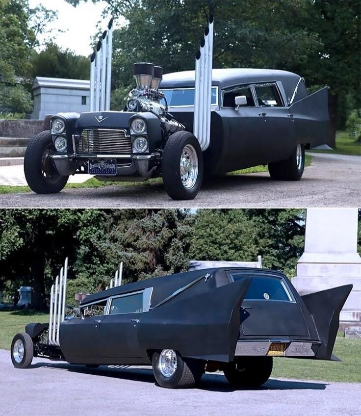 A highly customized black car, resembling a hearse or a stretched hot rod, with a prominent exposed engine and multiple tall, vertical exhaust pipes. The vehicle features large, bat-like fins at the rear. It is parked outdoors, possibly in a cemetery or park setting, with trees and a stone structure in the background.