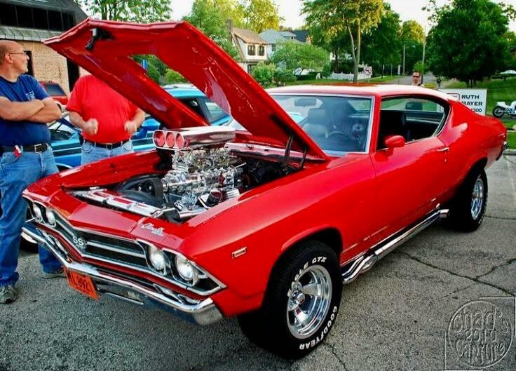A red classic muscle car, likely a Chevrolet Chevelle SS, with its hood open, showcasing a powerful, customized engine with chrome parts and multiple air intakes. Two men are standing nearby, admiring the vehicle. The car has 'SS' on its grille and 'GOODYEAR GT E' on its tires. A watermark 'chad's 2010 capture' is visible in the bottom right corner