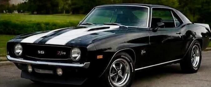 A classic black Chevrolet Camaro SS Z/28 with white racing stripes on the hood, parked outdoors with a green grassy background. The car features chrome bumpers and classic five-spoke wheels.