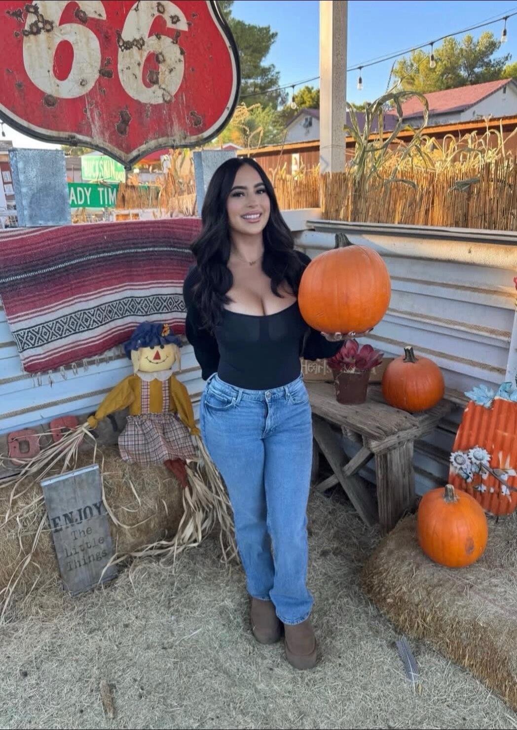 A woman standing at a pumpkin patch holding a pumpkin. There is a Route 66 sign in the background with fall decorations and hay bales.