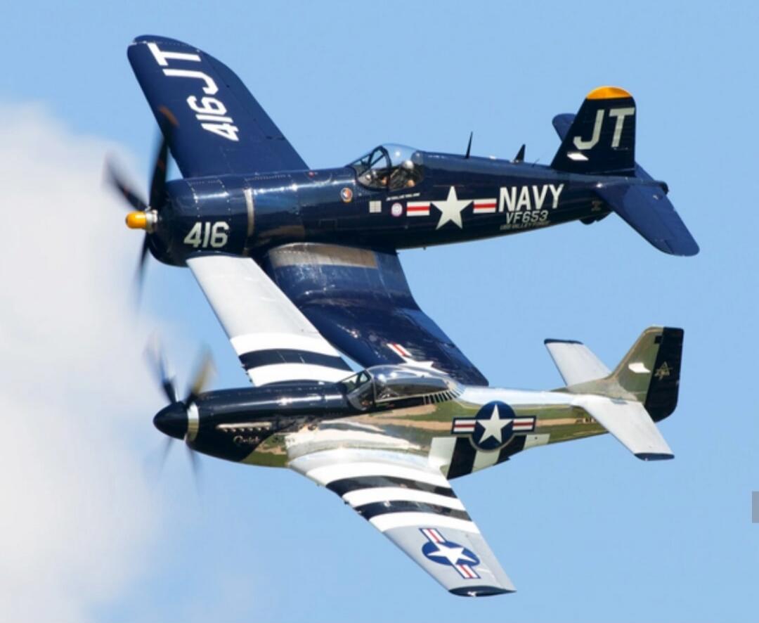 Two vintage warplanes, a dark blue F4U Corsair marked 'NAVY VF653 416 JT' and a silver P-51 Mustang, flying in a clear blue sky.